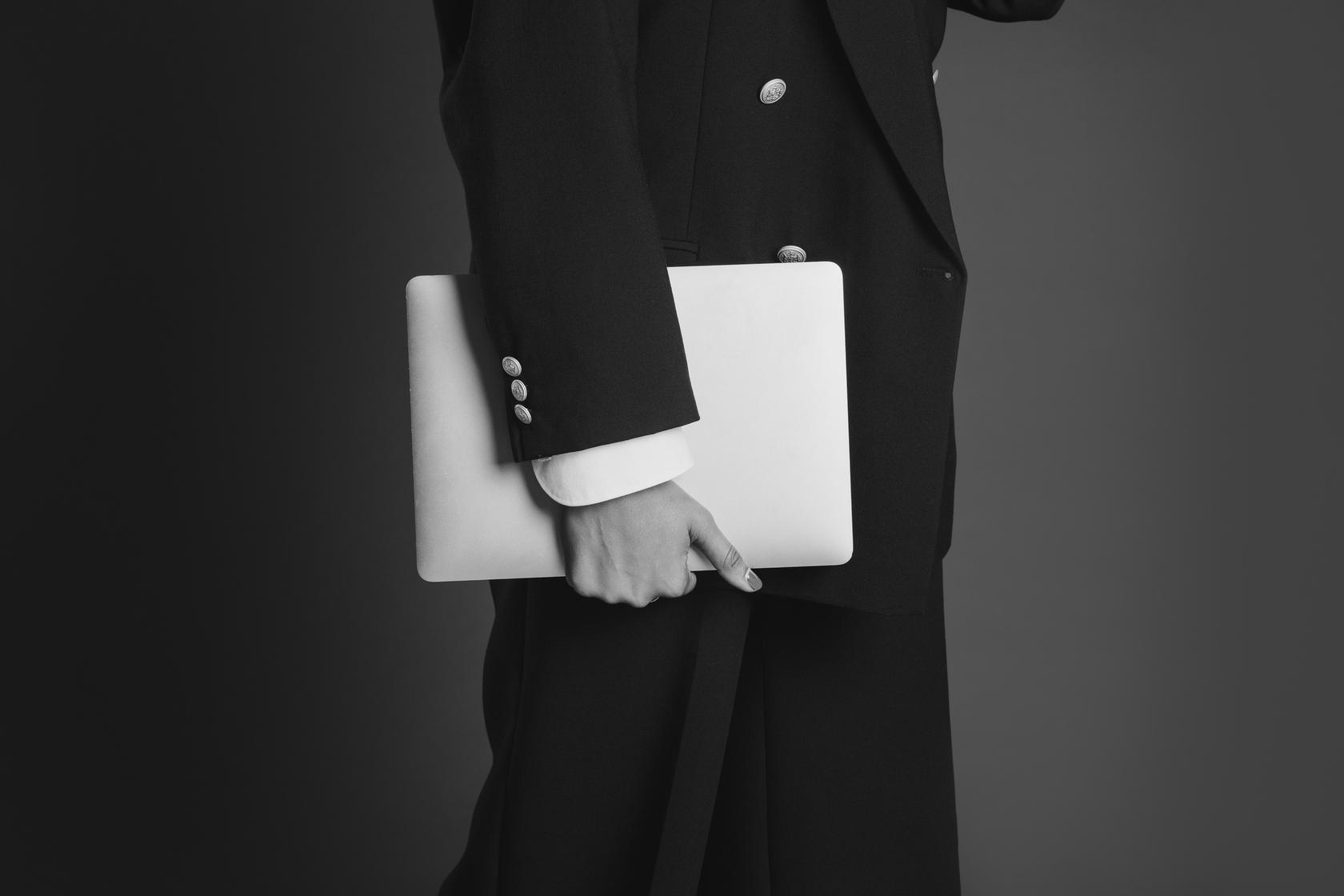 Mulher de blazer preto segurando um notebook junto ao corpo, fotografada do pescoço para baixo, em preto e branco.