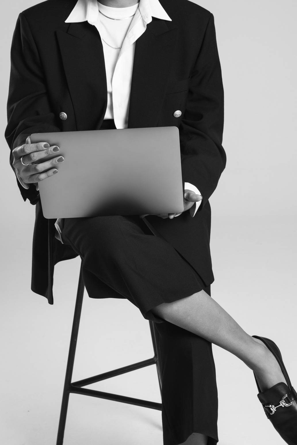 Mulher de blazer sentada em uma banqueta, com um notebook apoiado no colo, em preto e branco.