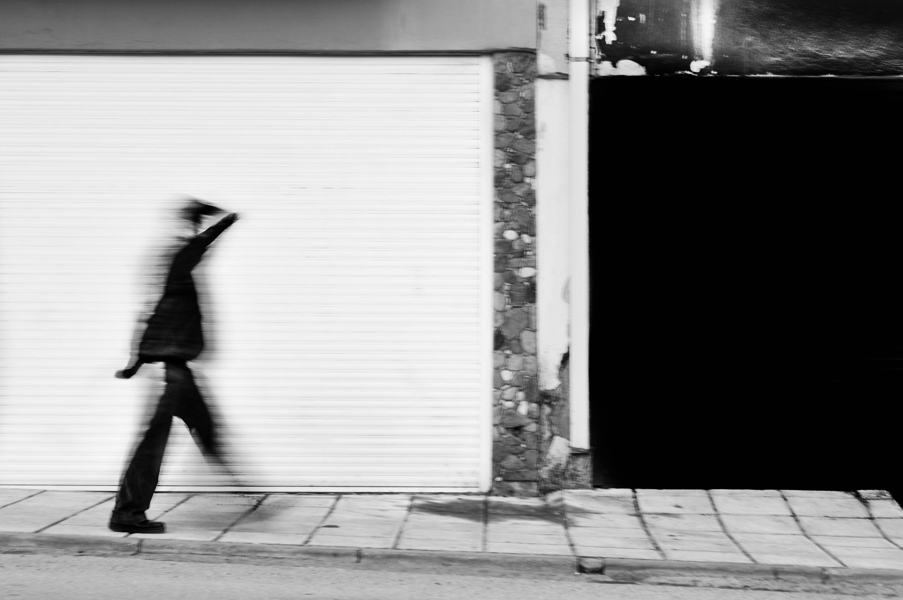Pessoa caminhando em movimento, desfocada, diante de uma porta de enrolar branca, em preto e branco.